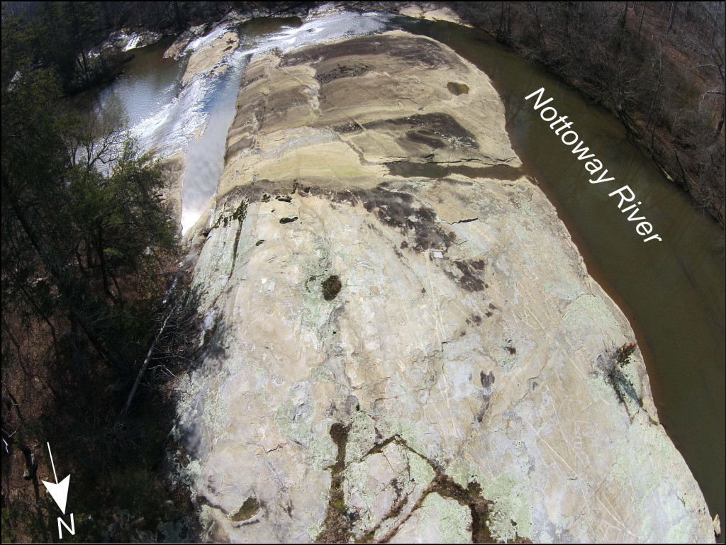 Oblique aerial view to the south of the uppermost outcrop at the Falls of the Nottoway, Virginia. At high water conditions, the Nottoway River flows over the top 1/3 of the outcrop, which is stained with a patina of iron and manganese oxides as well as a thin coating of sediment. In the upper part of the outcrop (the foreground) the granitic gneiss is rarely covered by water and the bedrock is better exposed. 