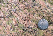 Outcrop photo of granitic gneiss in the Virginia Blue Ridge. K-feldspar is pink, quartz is blue-gray, and epidote is green. Note foliation from upper left to lower right and distinctive coin for scale.
