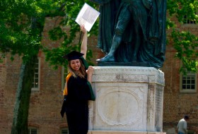 a graduating student holds up their degree next to the statue in front of the wren building