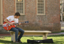 Male student playing guitar on a bench near the Wren Building