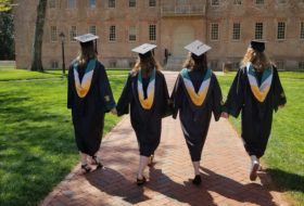 Four students wearing caps and gowns hold hands as they stroll down a brick path