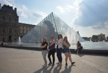 Four friends pose in front of the glass pyramid at the Louvre museum in Paris.