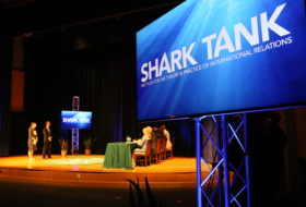 Shark Tank sign on a screen on a stage with people presenting in the background