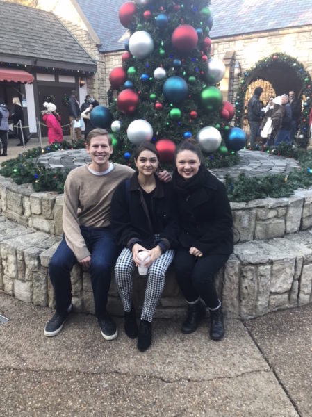 Three William & Mary students sitting on a bench under a decorated Christmas tree in Christmas Town at Bush Gardens. 