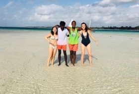 Four smiling people in bathing suits standing in the shallow waters of a white sand beach with arms around each other.