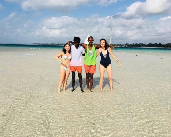 Four smiling people in bathing suits standing in the shallow waters of a white sand beach with arms around each other.