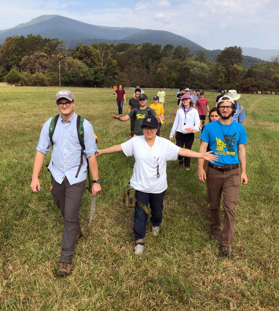Group of 19 students walking across a field in rugged clothing, smiling, with trees in the background and mountains further in the background.
