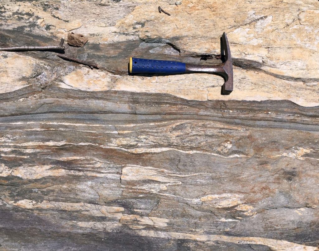 A hammer sits on the granite for scale. More lighter sample is at the top and below is mostly darker gray gneiss with some layers of the lighter granite layered in.