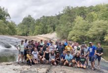 Thirty-four smiling participants group together in rugged shorts and tshirts for a photo on the gently sloping bedrock ledges of Nottoway Falls. Trees beyond the waterfall.