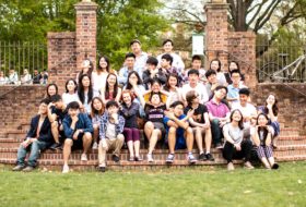 Group of approximately 30 students seated and posing for a group photo on the curved brick steps of the Sunken Garden near the Wren Building.