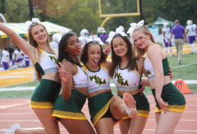 Five W&M Tribe cheer leaders posing for a photo next to the football field in uniform with a mix of silly faces and smiles.