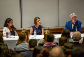 Three people sit at the front of a room at a long panel table, with microphones. Backs of audience members visible.