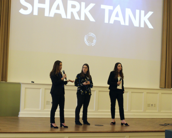Three students in business attire on stage with microphones presenting. Slide on large screen behind them reads Shark Tank