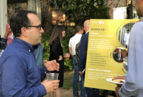 Faculty, staff and students in business casual attire, holding refreshments and discussing research posters on display in the tree-lined front yard of the GRI house.