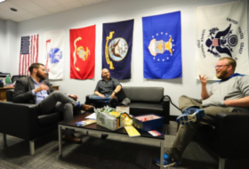 An office space with military flags on the wall and comfortable conversation chairs and couches with two students talking with Charlie Foster