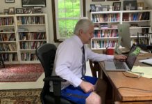 Professor Evans sitting at his desk on Zoom in front of a laptop wearing a shirt and tie, and casual shorts and shoes.