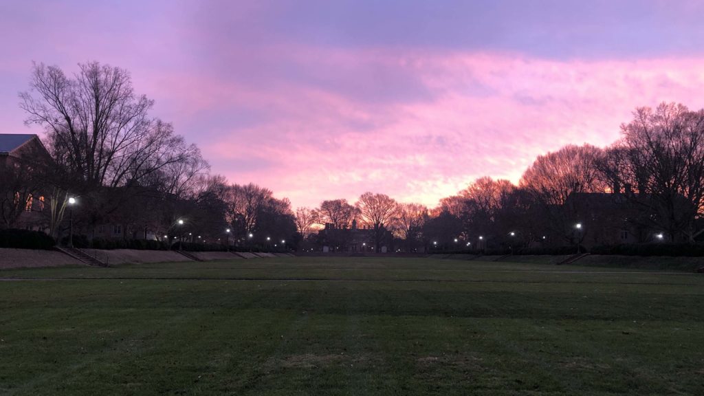 Sunrise over a wide, flat grassy area, sunken, with surrounding trees and academic buildings at William & Mary.