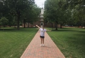 A person standing with arms outstretched joyfully on the brick pathway leading through grass and trees to the historic Wren Building on campus.