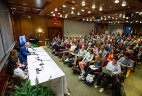 Three panelists sitting at a long table with microphones, and a primarily student audience seated in rows of chairs.