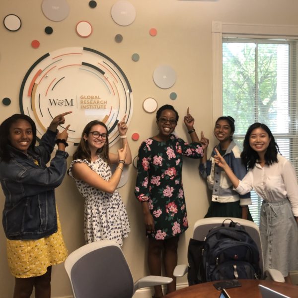 Group of five students smiling and pointing to a collage installation of circles on a wall, surrounding the Global Research Institute's circular logo.