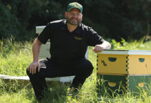 Person sitting in a field next to a pair of green and gold bee hive boxes with the W&M logo on them.