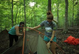 Two masked campers pitching their tent in the green woods of campus.