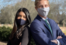 Two masked people, posed back to back with their arms crossed, in professional attire.