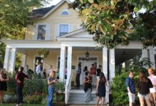 Two story yellow house with a large front porch with clusters of students and faculty chatting together