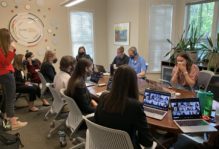 A group of 11 masked people around a conference table with laptops open with Zoom meetings in progress.