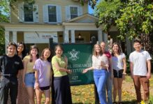 Nine smiling students pose with the Global Research Institute sign in the front yard of a yellow house.