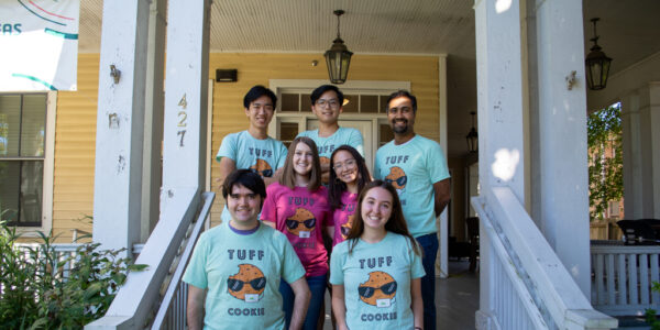 Seven smiling people wearing TUFF Cookie tshirts grouped on the porch of a large yellow house