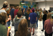 A classroom of masked people standing in groups facing the front of the room