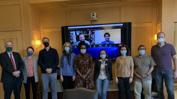 Nine masked people pose together in front of a large monitor displaying two participants on Zoom.