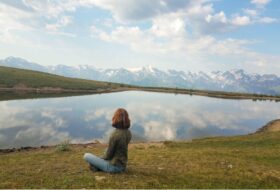 A person sitting beside a mirrored lake reflecting blue skies and white clouds, with snow-capped mountains in the distance.