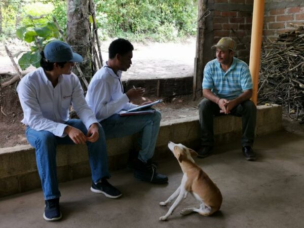 Three people sitting in an outdoor structure and talking. One person is holding a pen and clipboard and a dog is sitting at their feet.