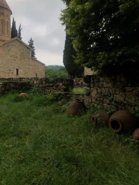 Large pottery vessels tipped in the grass against a low stone wall with a stone building and tall trees in the background