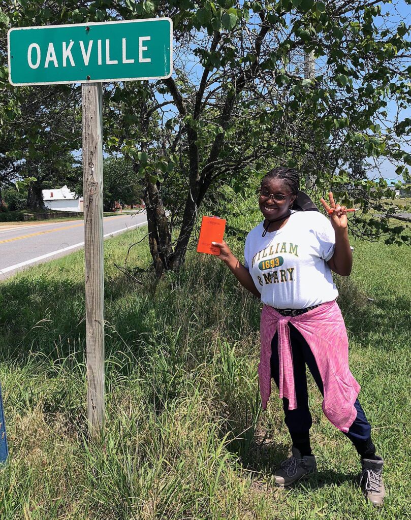 A person in a W&M t-shirt smiling next to an Oakville sign on the road side.
