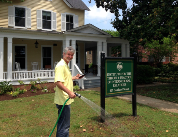 A smiling Mike Tierney holding a water hose in front of a large yellow house with a sign that reads: Institute for the Theory & Practice of International Relations, 427 Scotland Street