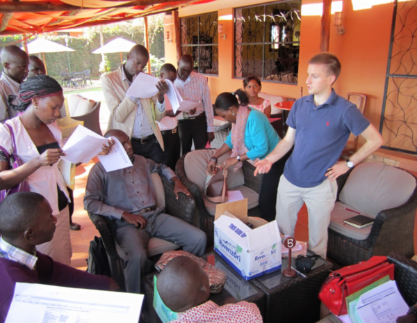 A covered work area with a dozen people reading materials and discussing with Austin Strange in Uganda.