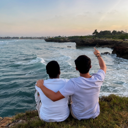 View from behind two people sitting on the edge of a body of water, overlooking the lapping blue water and pointing at the sunset