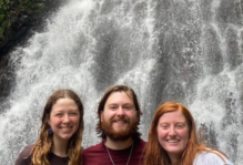 Three arm-in-arm smiling people in front of a rushing waterfall
