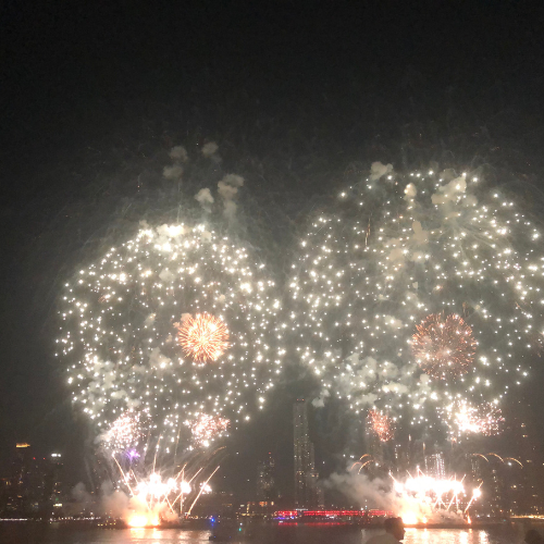 Two large fireworks displays above barges on a river with skyscrapers in the background