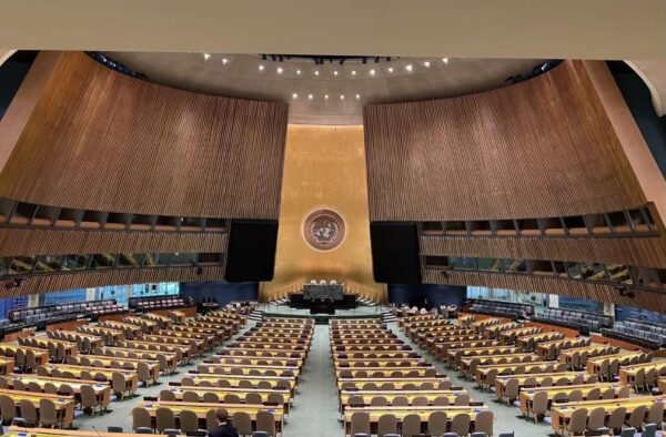 The large United Nations hall with many rows of empty tables and chairs facing the large dais and podium at the front.