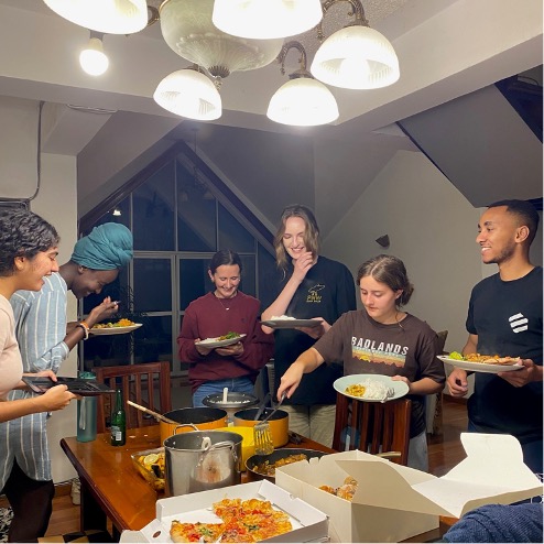 Six people smiling and laughing while serving up their plates with a variety of homecooked and takeout food.