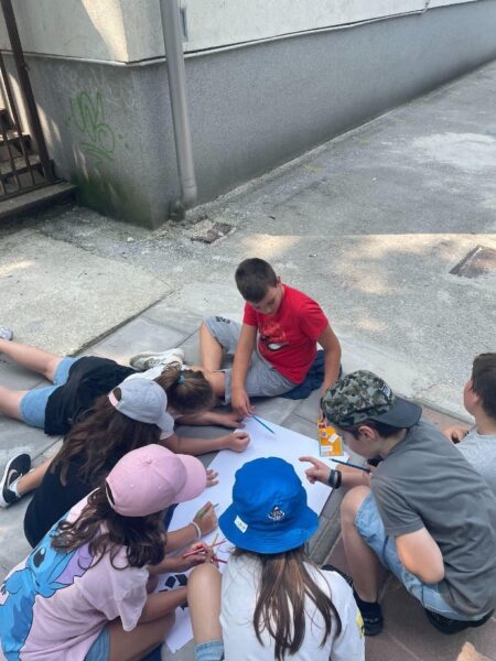 A group of kids seated in a circle around a poster board on the sidewalk, all drawing on it