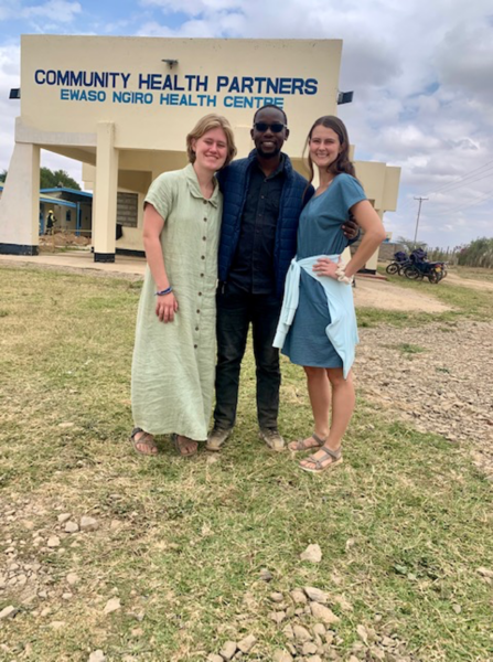 Three smiling people posed in front of a low building with Community Health Partners, Ewaso Ngiro Health Centers in blue lettering
